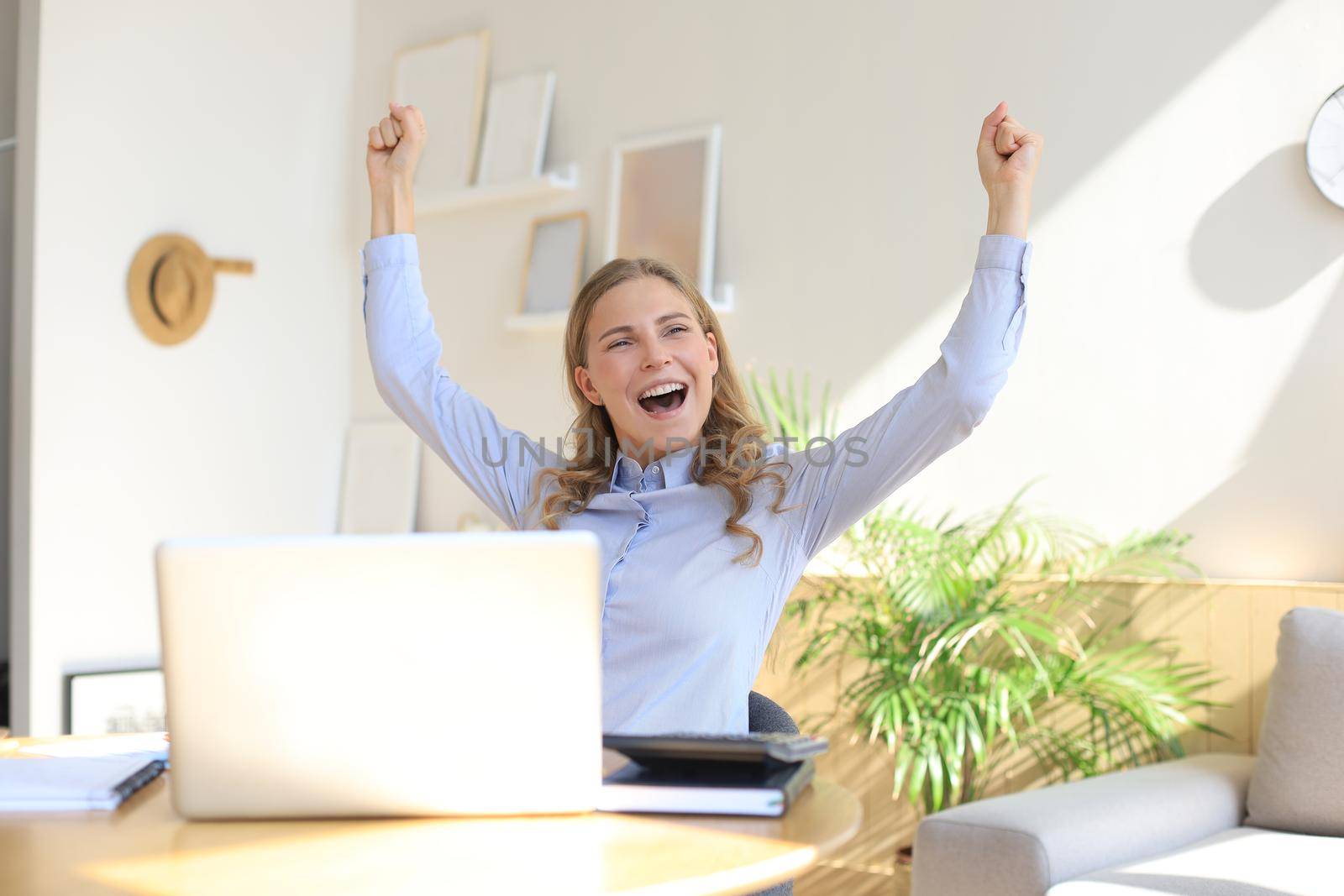 Happy entrepreneur woman sit at desk reading good news and express joy rising hands up. by tsyhun