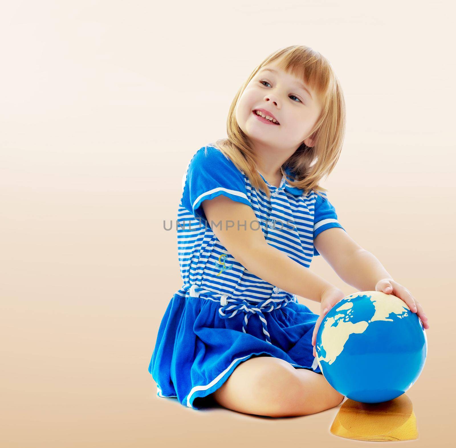 On a brown background a smooth transition from dark to light. Nice little girl in a Sea blue dress sitting on the floor. Girl turns hand the globe.