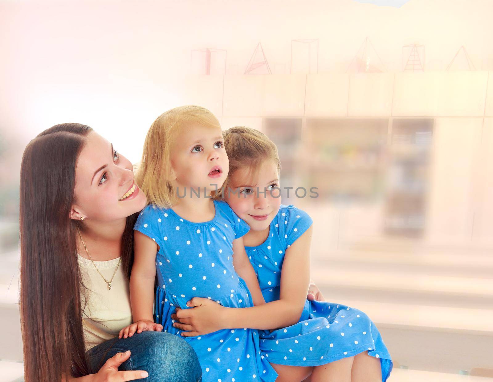 Beautiful young mother with her two daughters. All look up.On the background of the room where children live, and are on the shelves of toys.1
