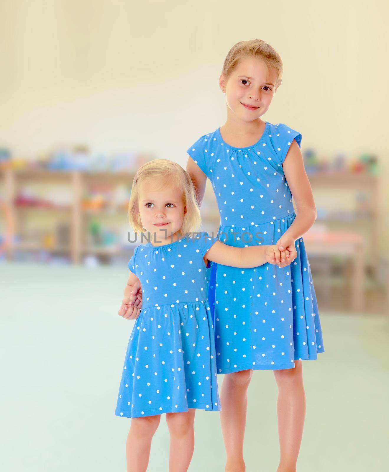 Against the background of a child's room .Two charming little girls, sisters , in identical blue dresses with polka dots , cuddling.