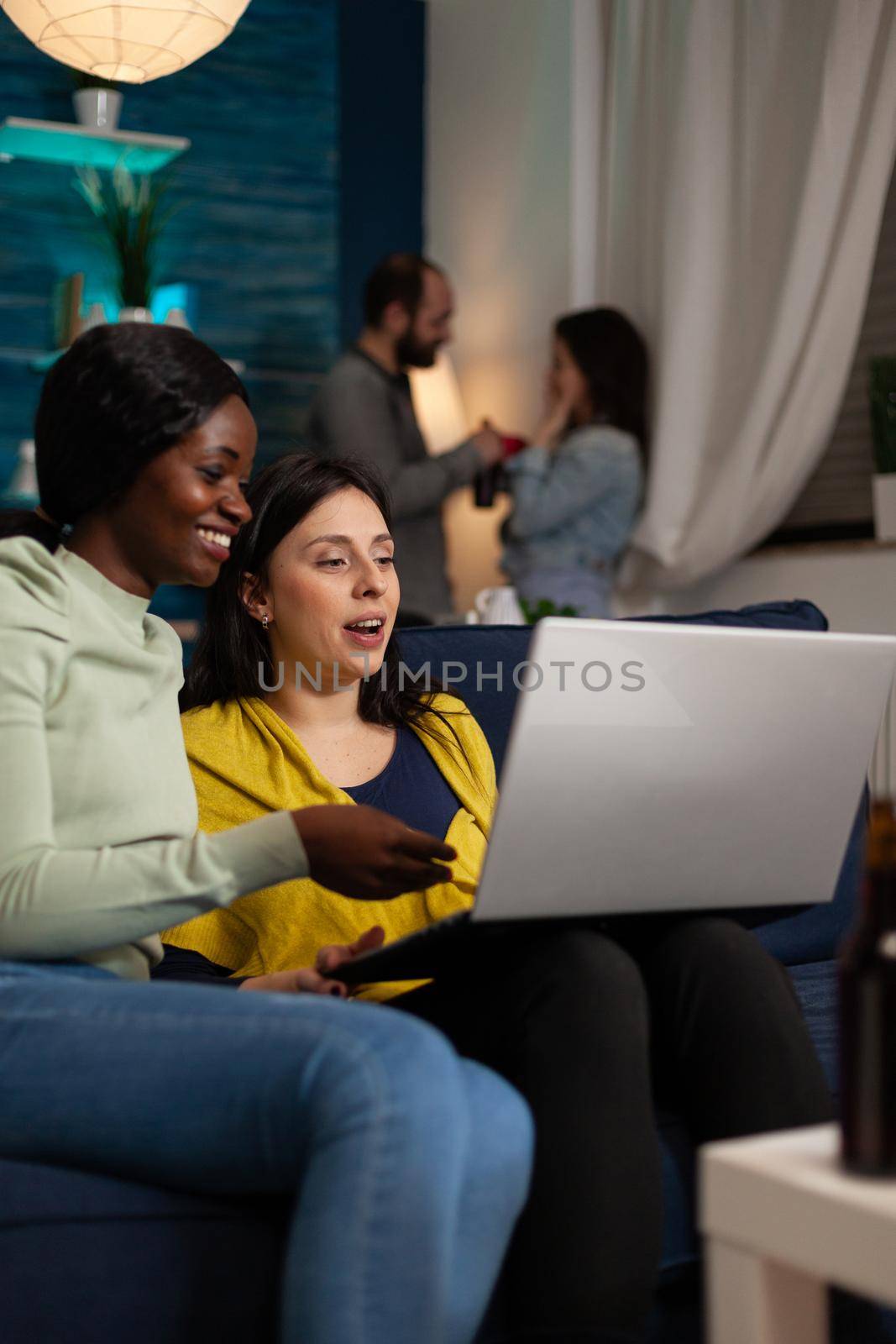 Smiling women looking at funny video on internet holding laptop computer sitting on sofa enjoying spending time together. Group of people hanging out during wekeend party at home. Friendship concept