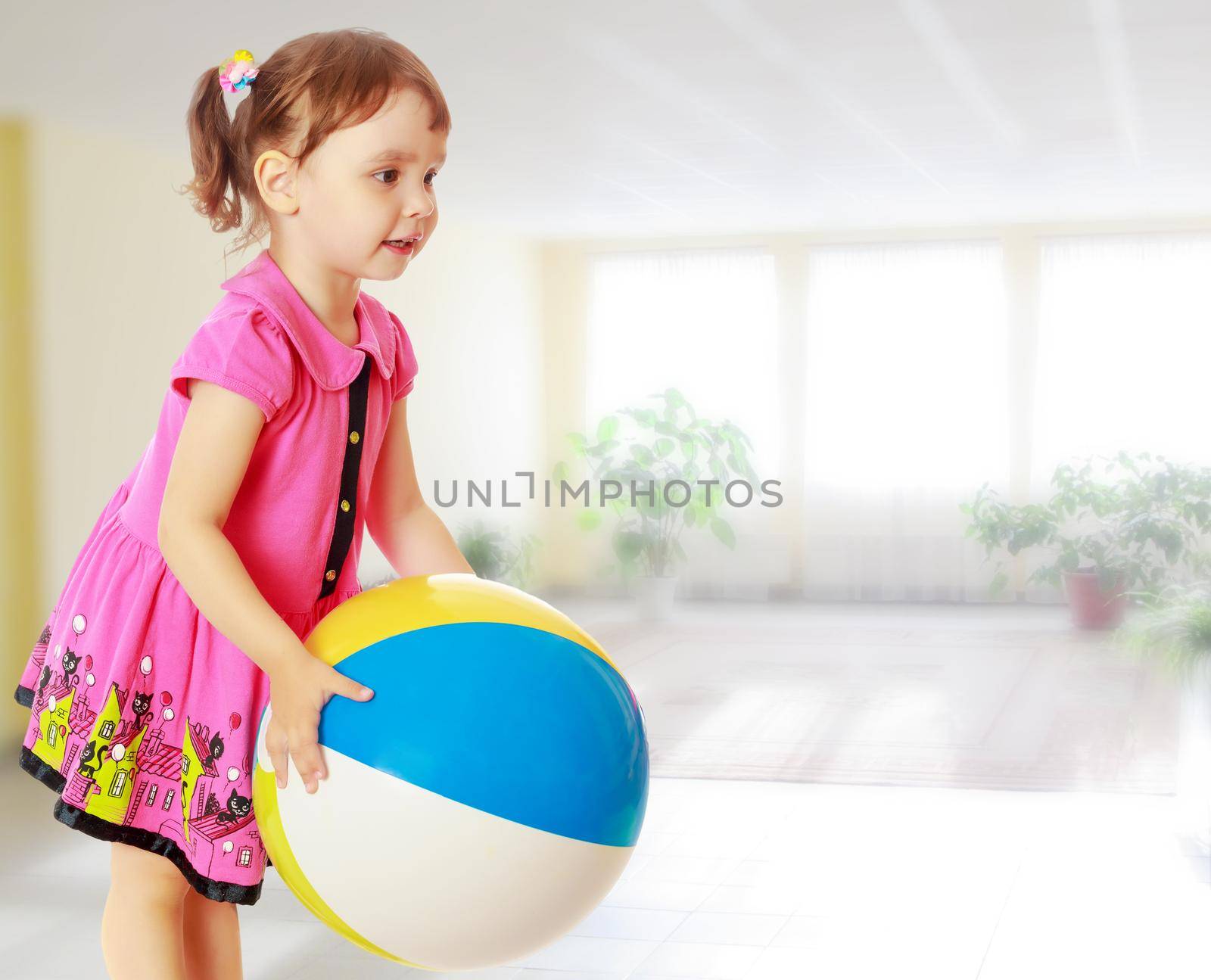 Beautiful little girl in a pink dress throwing a big striped ball. Turned sideways. Close-up.Against the background of a large room with Windows and flowers .