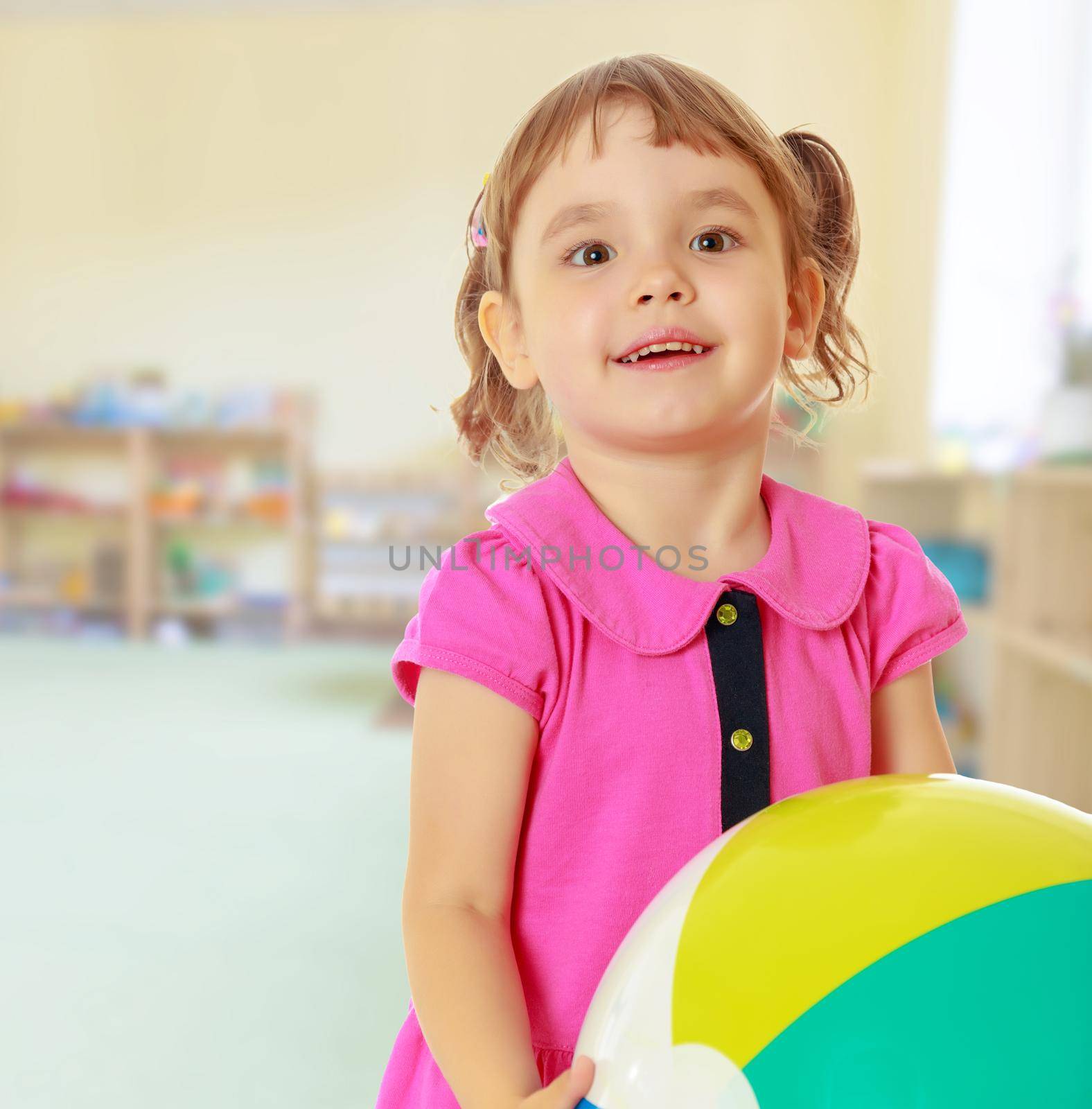 Cute little girl with pigtails on the head , in a pink dress. Girl holding a ball. Close-up.On blurred background the great hall of the kindergarten, with long racks where there are toys.