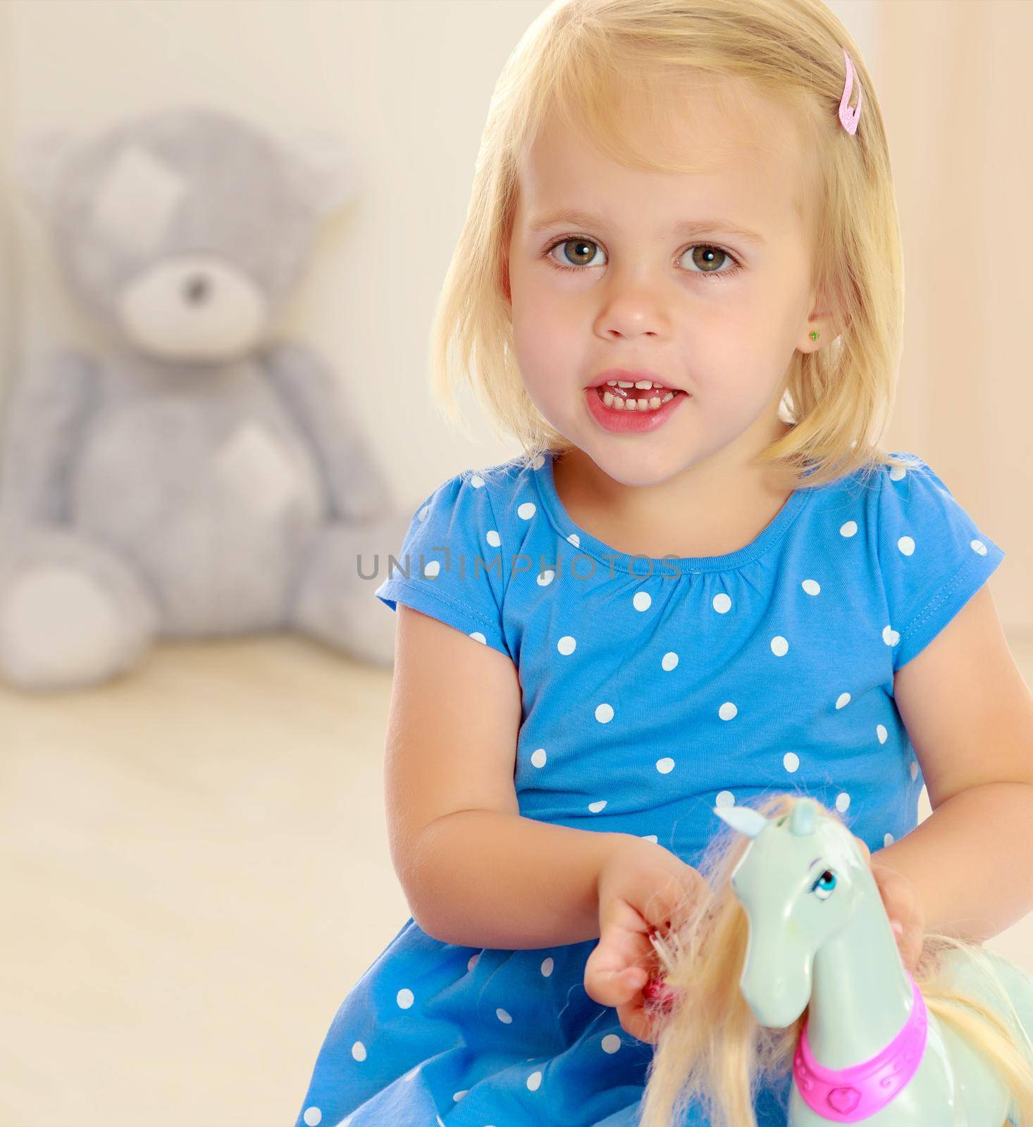 Cute little blonde girl playing with a toy horse. Girl wearing a blue dress with polka dots.In the background children's room where the sitting Teddy bear.