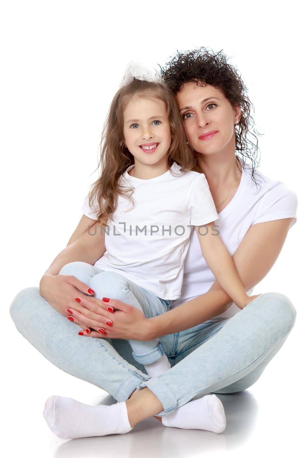 Beautiful, charming young mother with adorable little daughter. In the same jeans and white t-shirts without a pattern. Mom sits on floor and holds daughter on hands - Isolated on white background