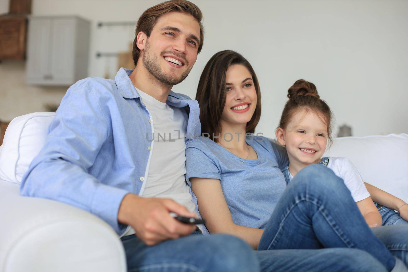 Happy family with child sitting on sofa watching tv, young parents embracing daughter relaxing on couch together. by tsyhun