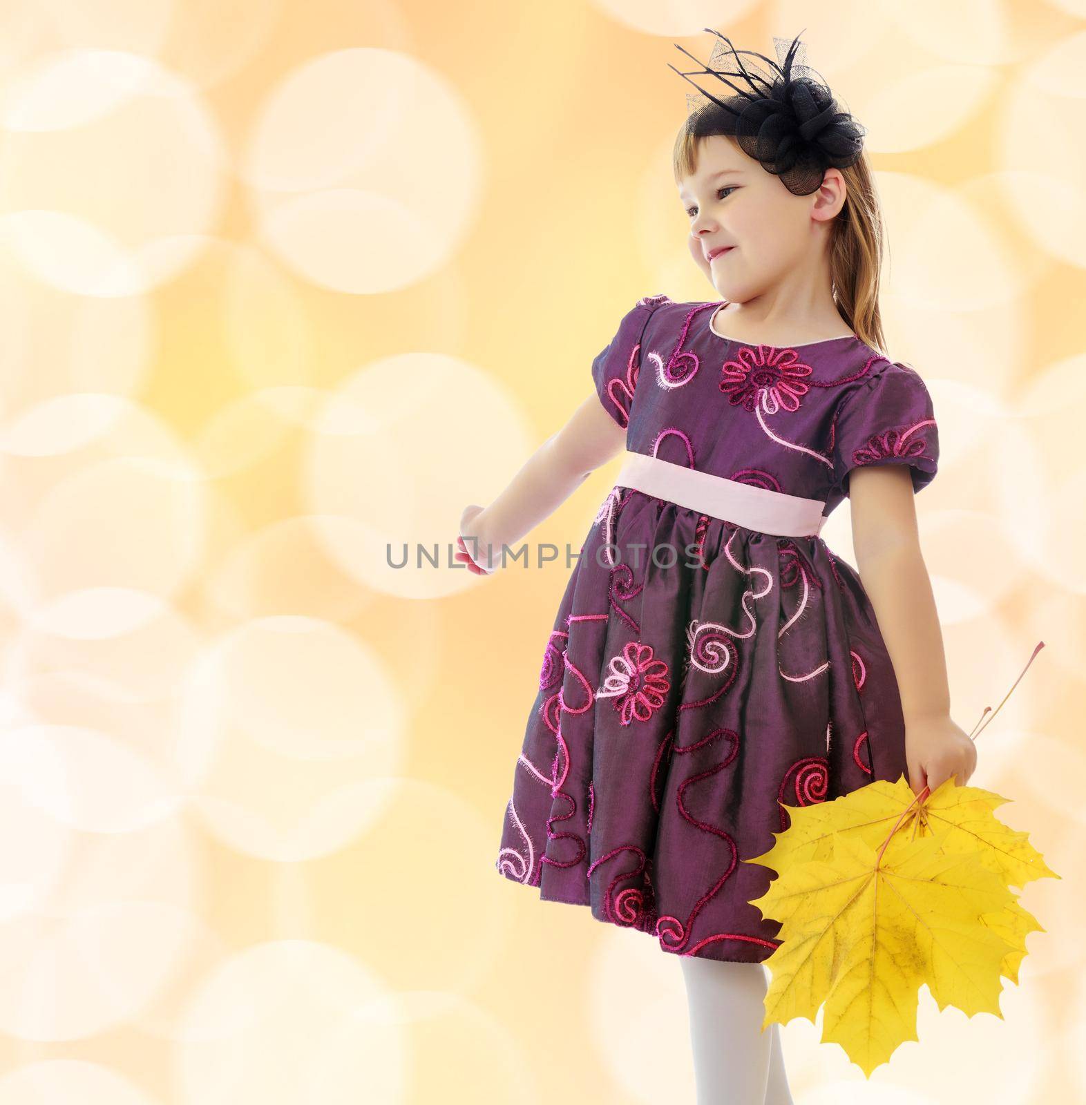 Caucasian little girl dressed in brown dress. She is holding a bouquet of maple leaves. Turning sideways to the camera.On a brown background with white blurred circles.