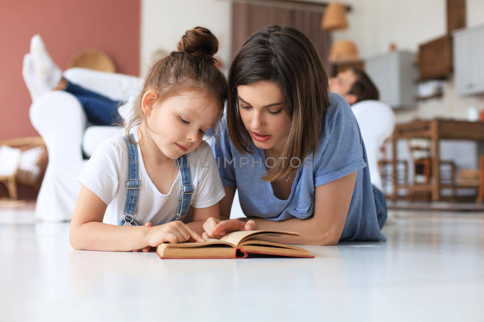 Mother and little daughter read a book together, lying on the floor in the living room