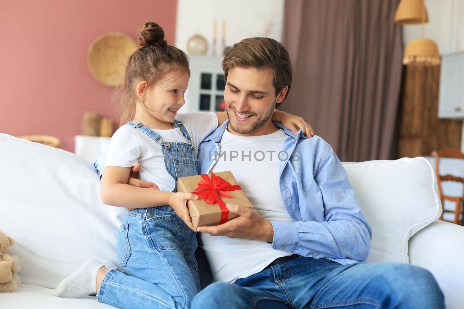 Cute kid daughter make surprise to daddy, little girl presents gift box to father sit on sofa. Father's day