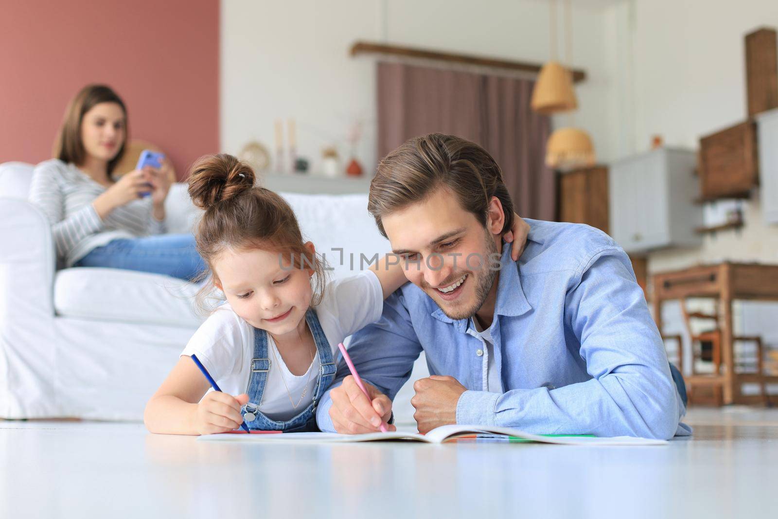 Happy father smilling daughter lying on warm floor enjoying creative activity, drawing pencils coloring pictures in albums, mother resting on couch, family spend free time together. by tsyhun