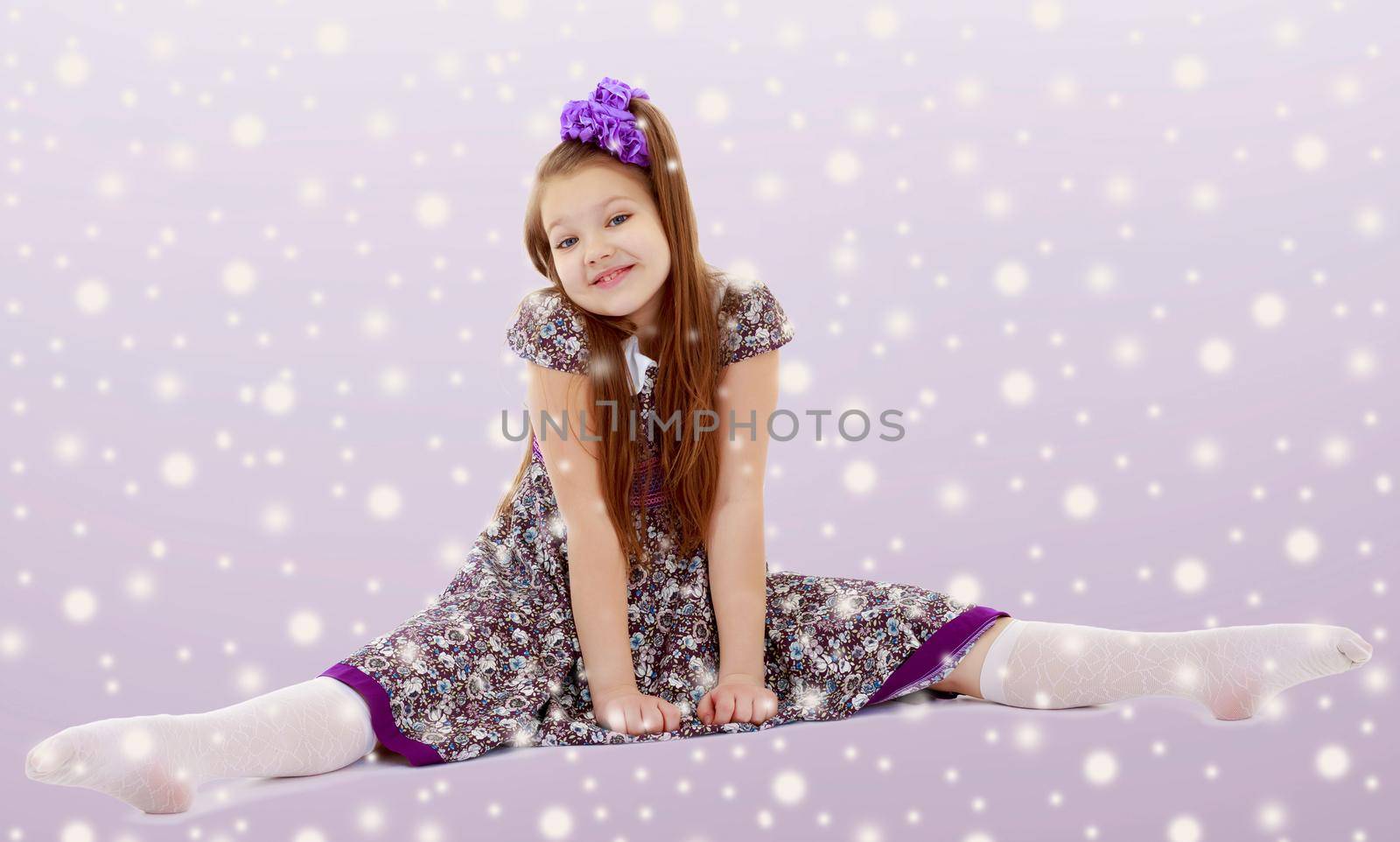 On the Christmas background with white snowflakes.Caucasian little girl with a big purple bow on her head. Girl shows how to do the splits.