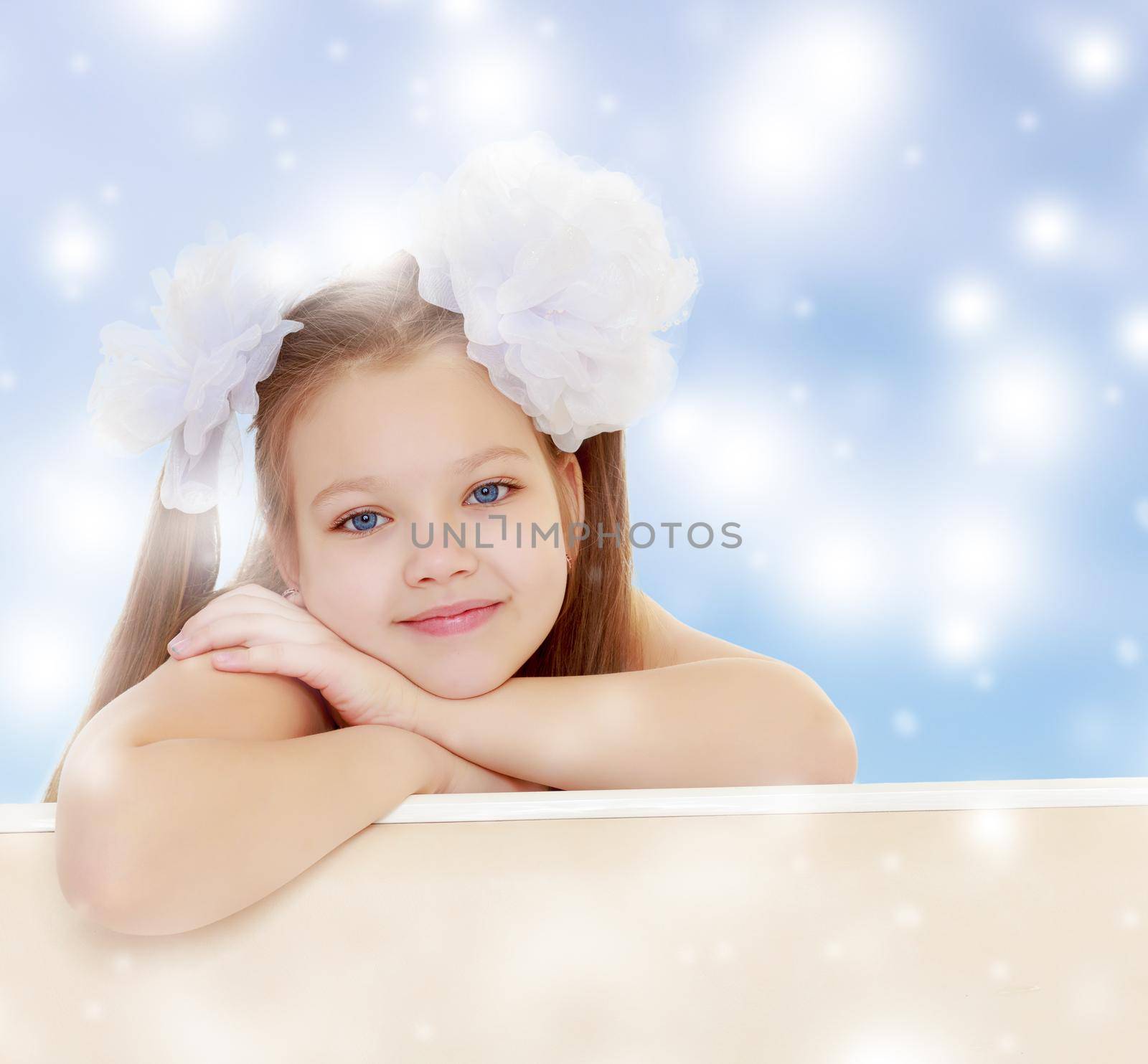 Gentle long-haired little girl with big white bows on the head . She leaned on the windowsill. Close-up.Blue Christmas background with white snowflakes.
