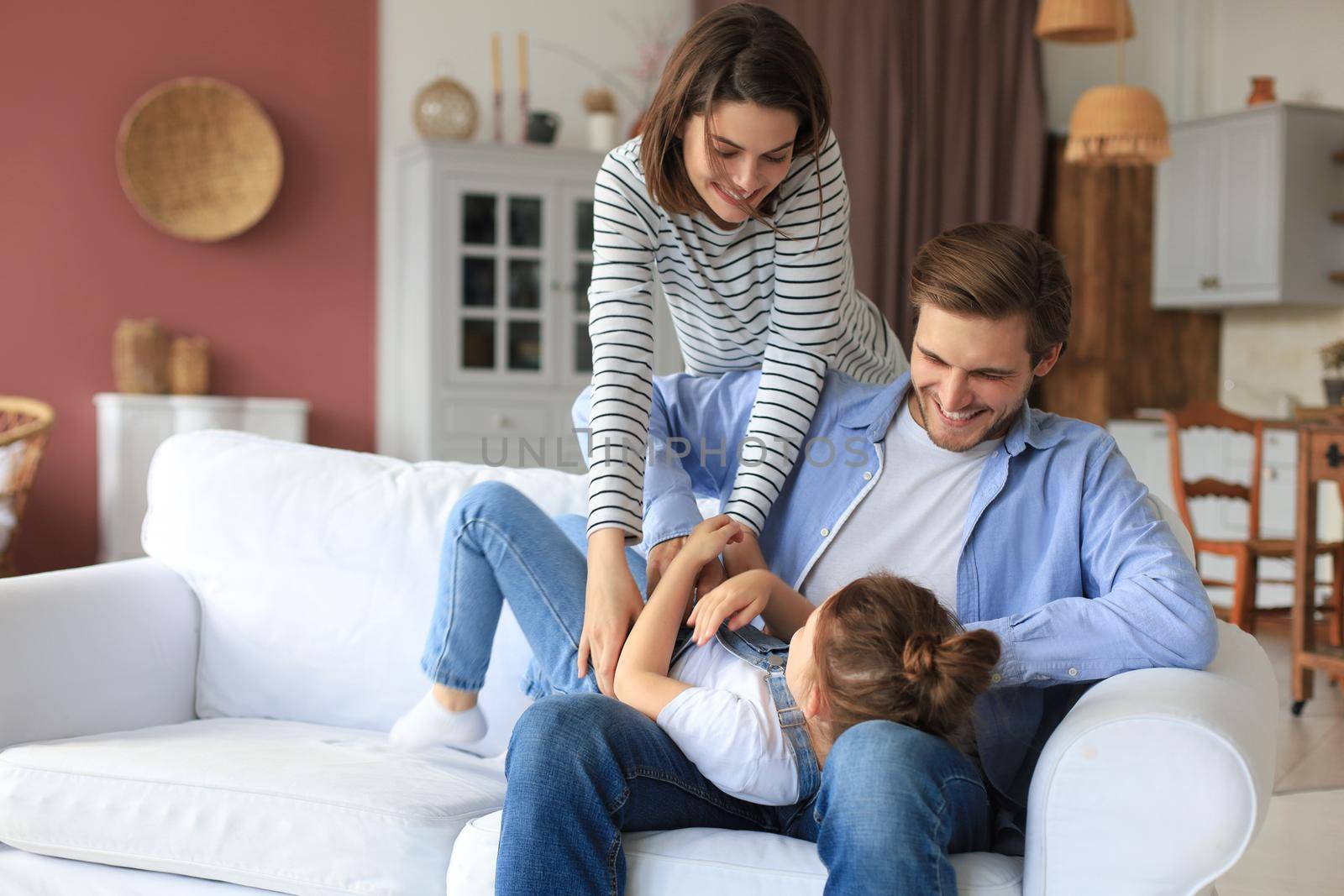 Positive friendly young parents with smiling little daughter sitting on sofa and having fun together at home