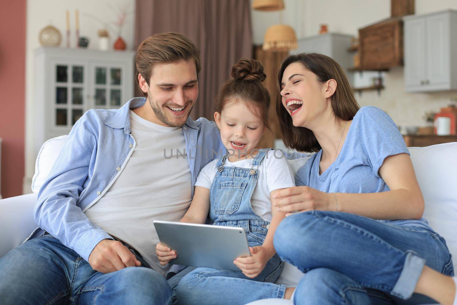 Positive friendly young parents with smiling little daughter sitting on sofa together answering video call on digital tablet while relaxing at home on weekend
