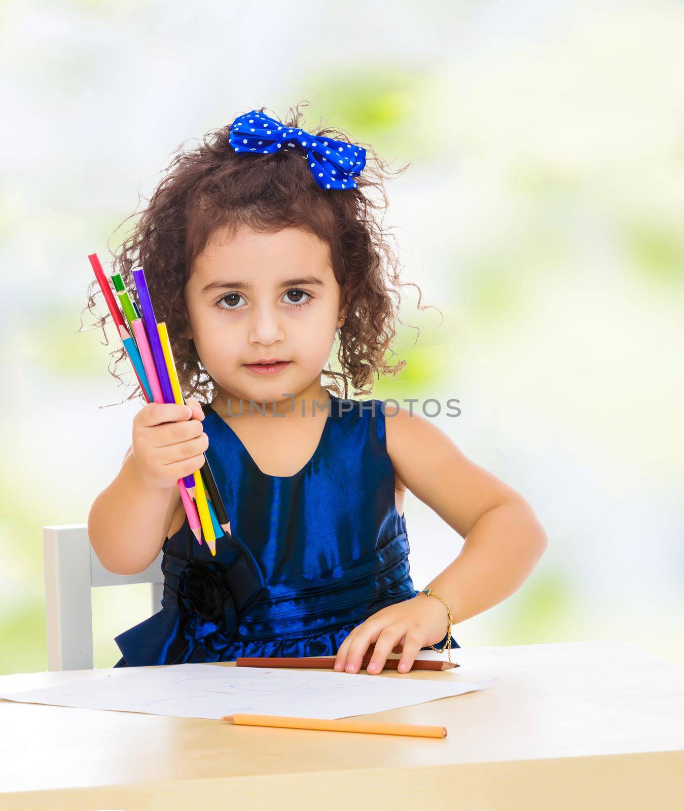 Cute little girl in blue dress, holds a lot of pencils . She paints at a table in a Montessori kindergarten.white-green blurred abstract background with snowflakes.