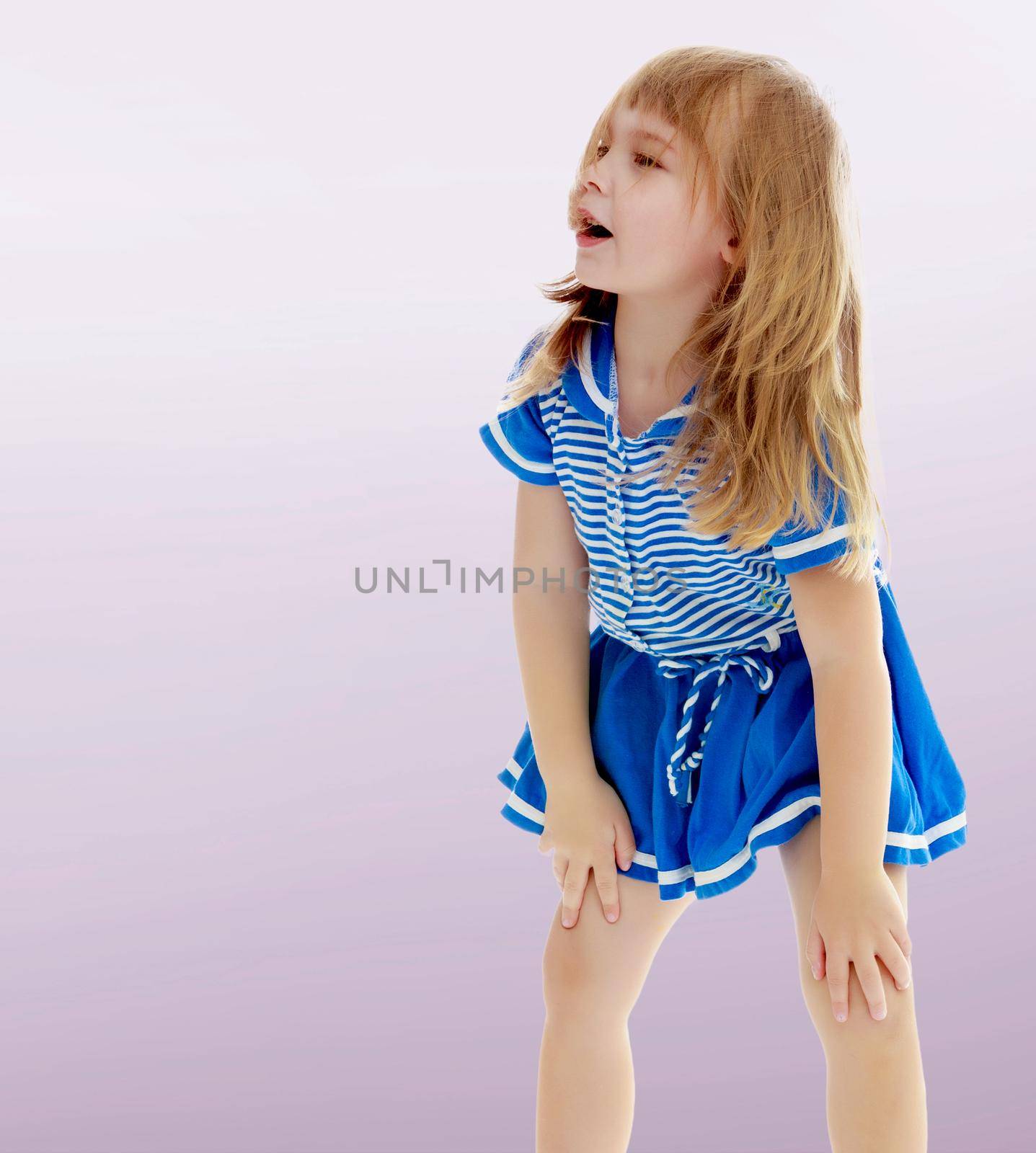 On a purple background, smooth transition from dark to light. Cute little unkempt girl in a short blue dress. Girl looking to the side with his hands on his knees.