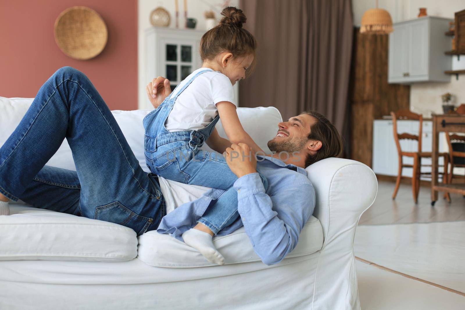 Smiling young father lying on couch at living room and play with happy little daughter
