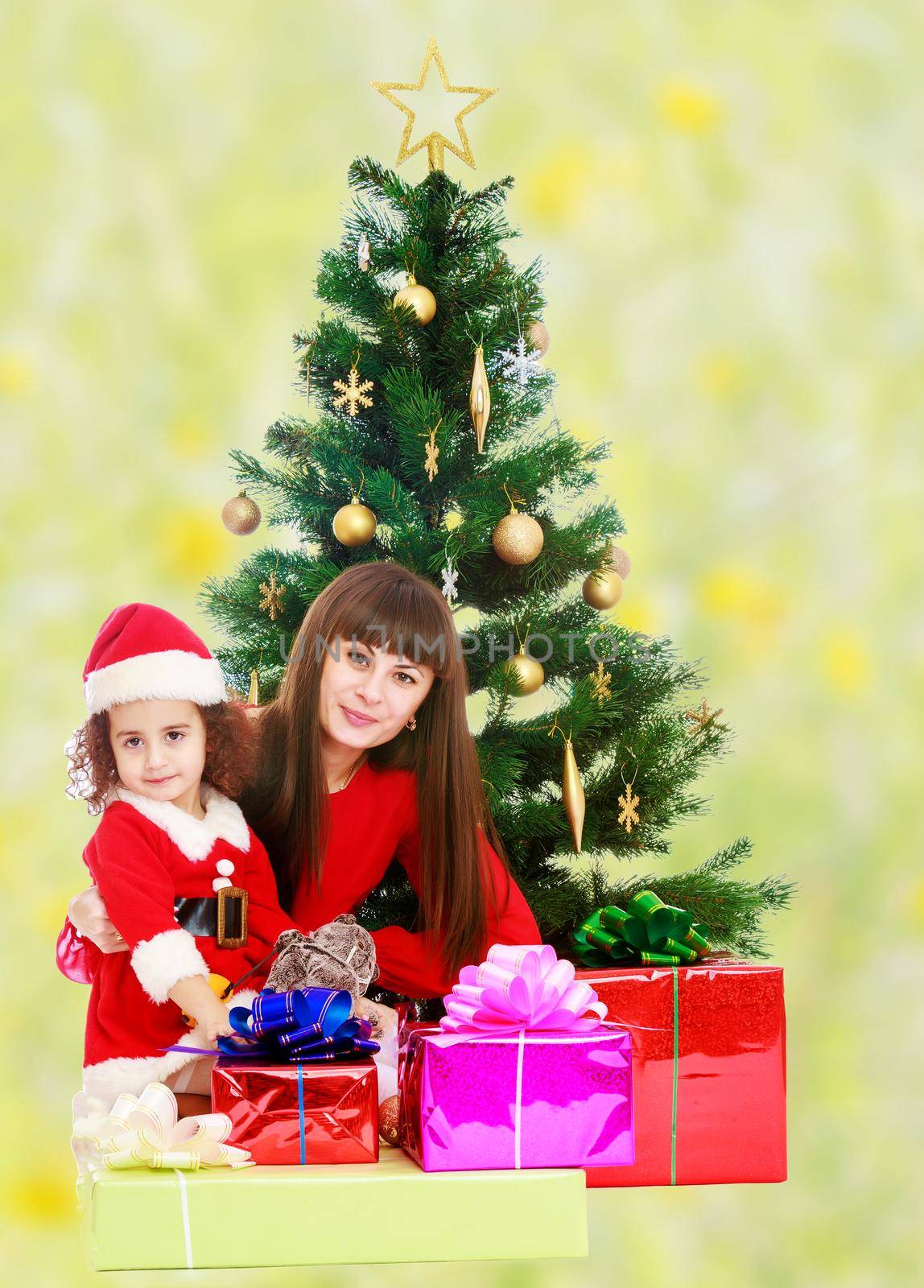 Beautiful mother and daughter , dressed in the costume of Santa Claus around the Christmas tree surrounded by heaps of gifts.Bright,floral yellow-green blurred background.