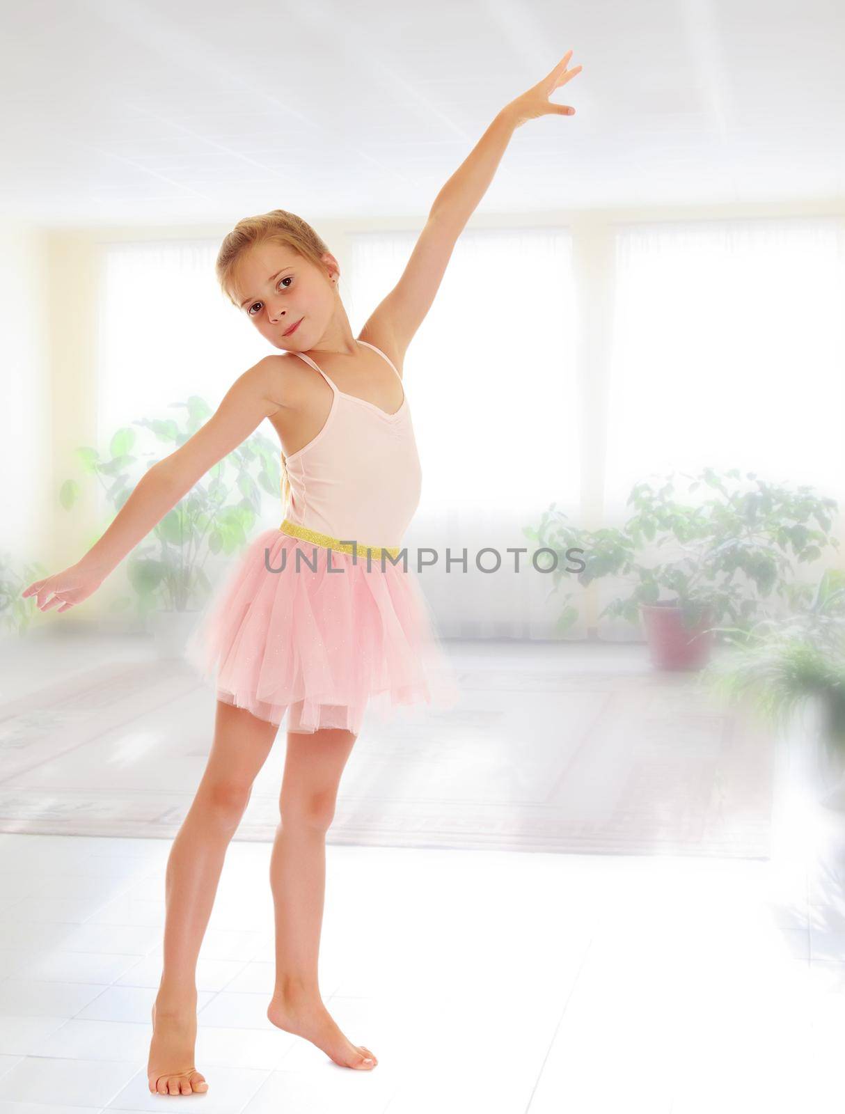 Charming little girl ballerina in a pink translucent dress.On the background of the school hall with large Windows.