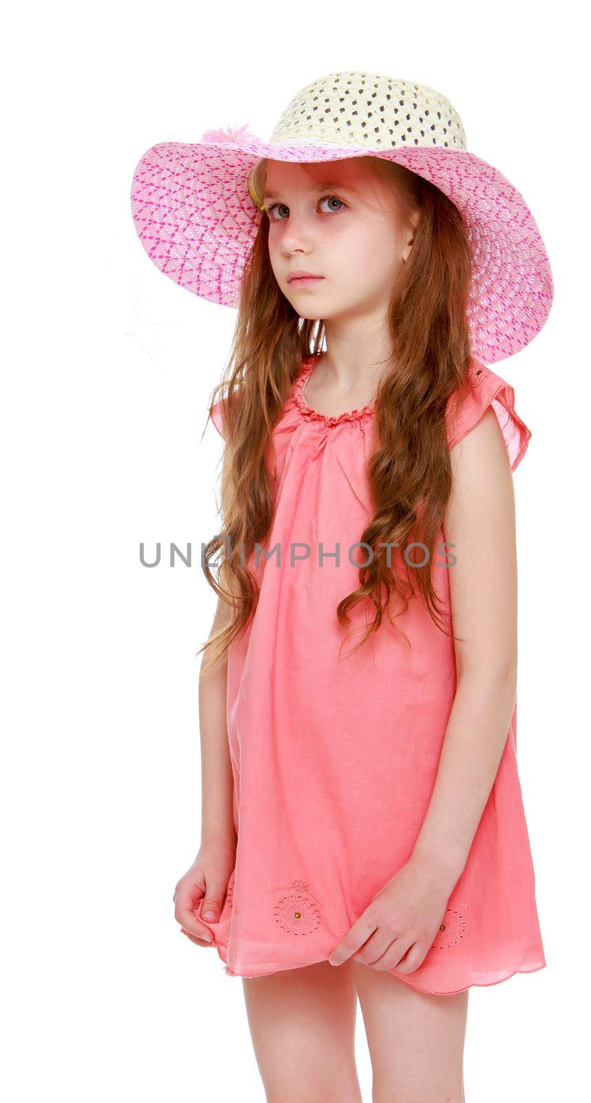 The doubting little girl with long pigtails to her waist and braided white bows. In short pink summer dress and pink hat. Standing sideways to the camera. Close-up - Isolated on white background