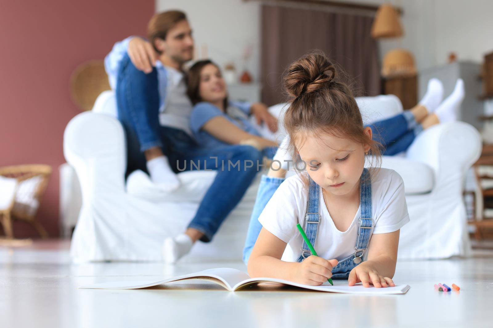 Smiling happy family sit relax on couch in living room watch little daughter drawing in album with colorful pencils. Happy weekends at home