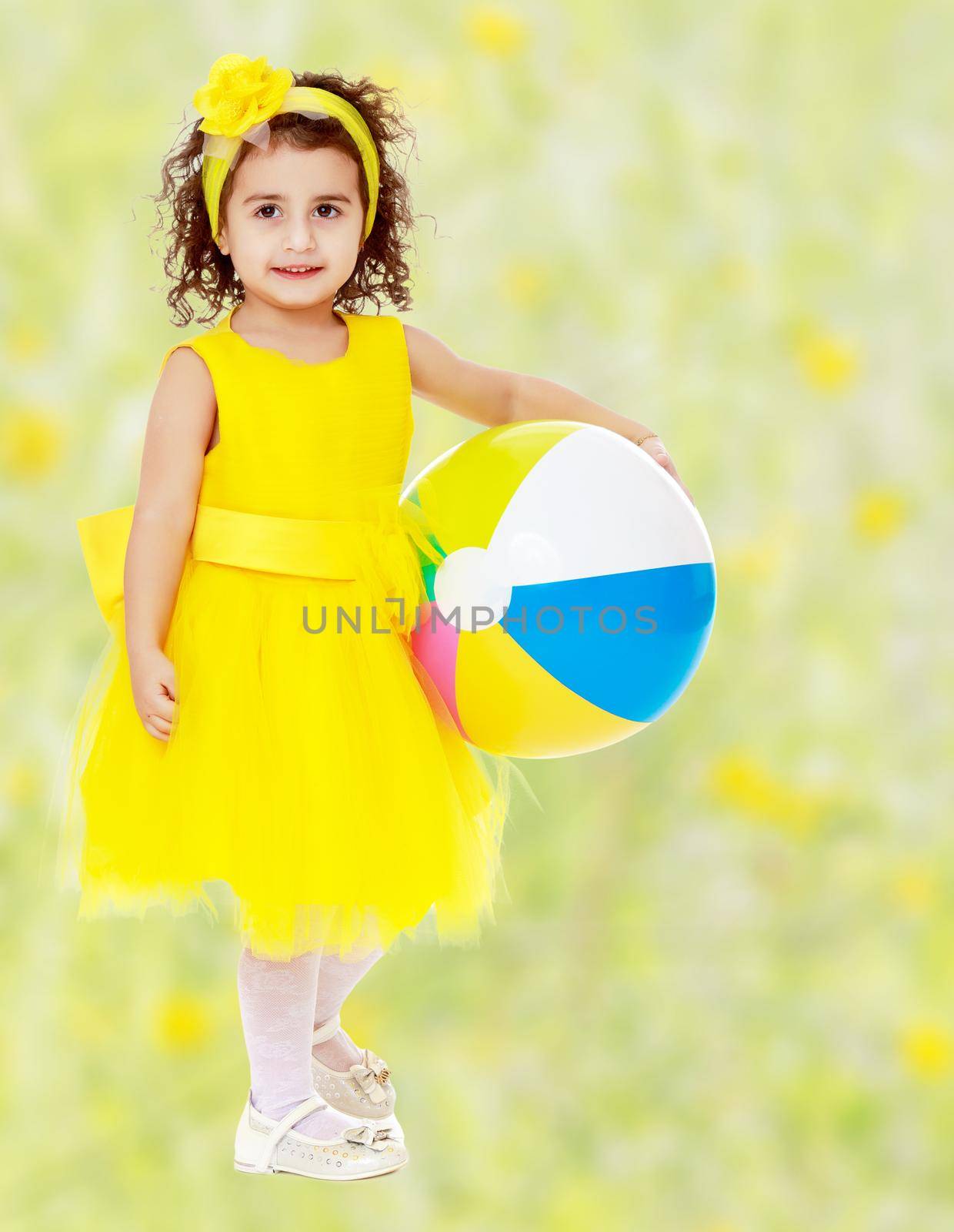 Cute curly girl in a bright yellow dress and bow on her head holding the ball. Close-up.Bright,floral yellow-green blurred background.