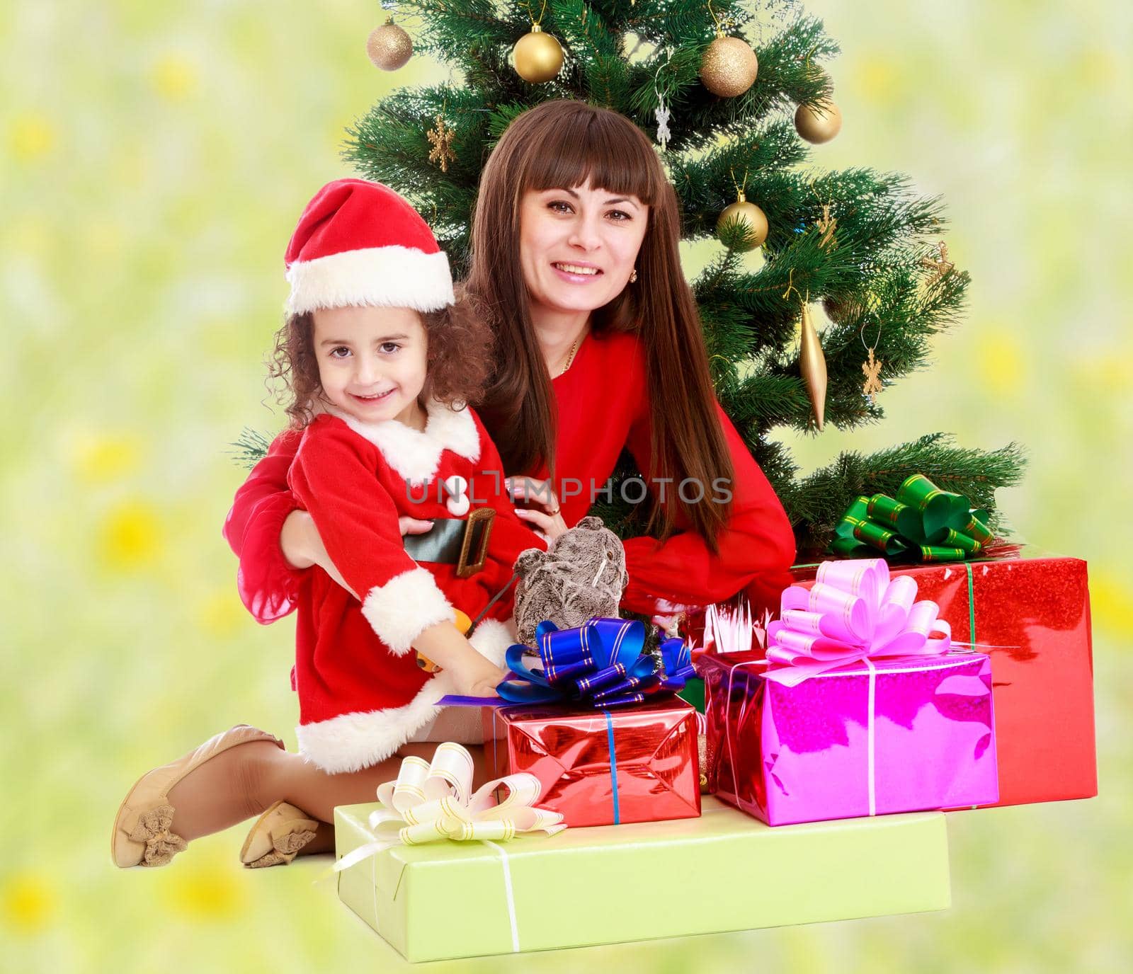 Happy mother and daughter near a Christmas tree surrounded by heaps of gifts.Bright,floral yellow-green blurred background.