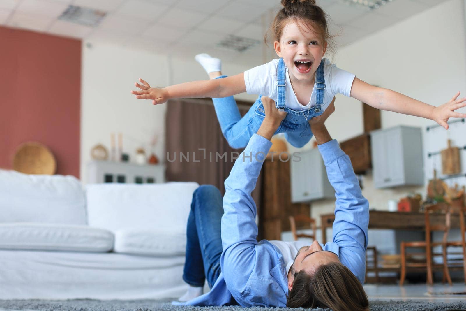 Loving young father lying on floor at home and play with laughing little daughter throws up in air like airplane