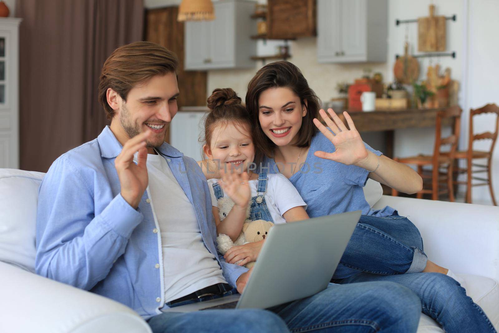 Positive friendly young parents with smiling little daughter sitting on sofa together answering video call on laptop and waving hand in greeting while relaxing at home on weekend