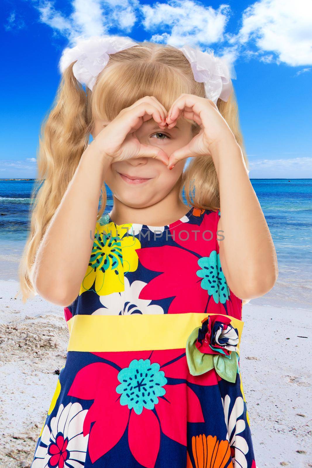 Beautiful little girl with long blond hair braided in ponytails , folded palms heart.On the background of sea beach, warm sea and blue sky with clouds.