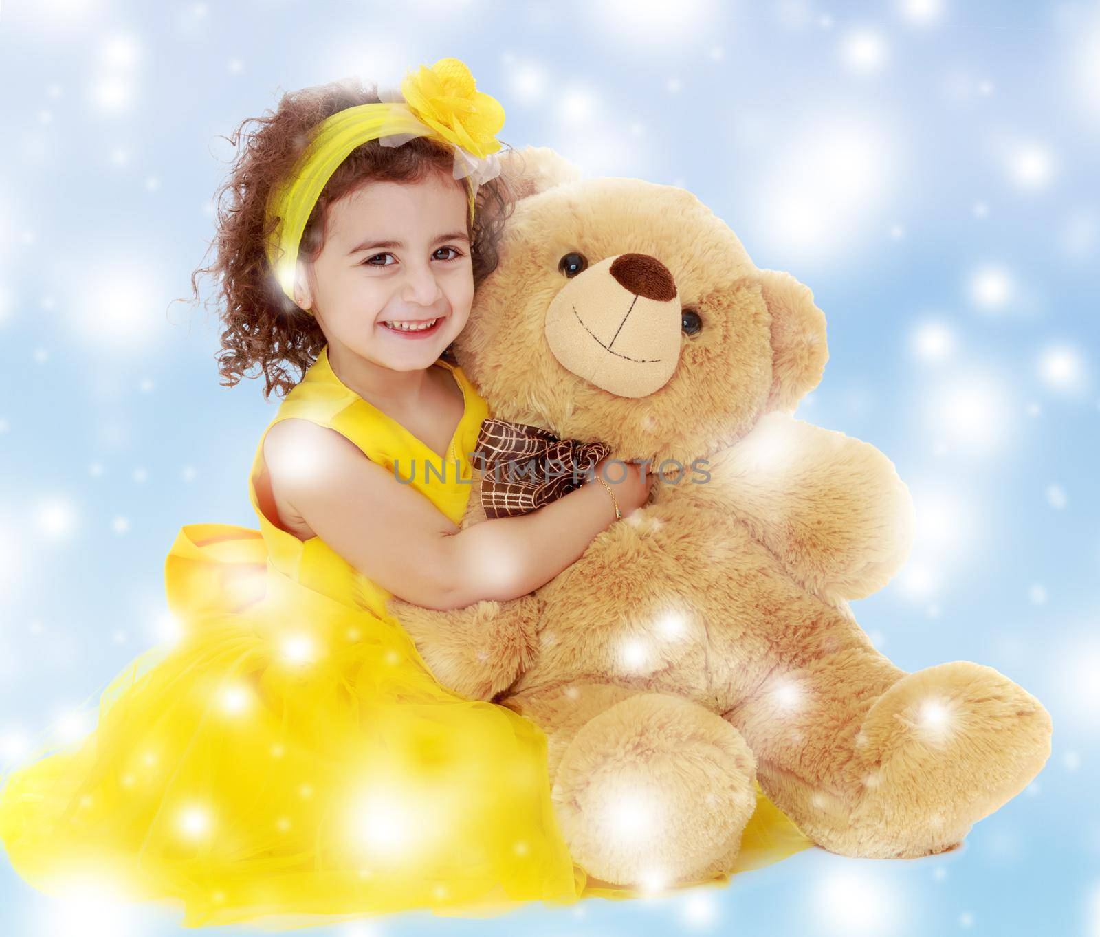 Joyful little girl in a yellow dress and bow on her head sitting on the floor. Girl hugging a big Teddy bear.Blue winter background with white snowflakes.