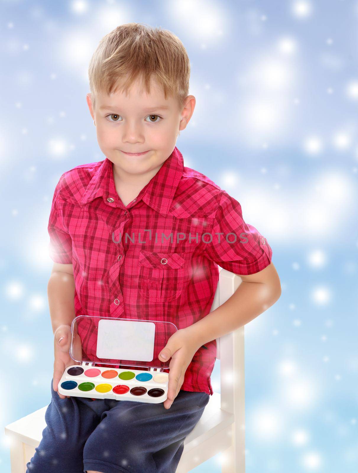 Cute little boy in a red shirt holds his box of watercolors.The concept of celebrating the New year, Holy Christmas, or child's birthday on a blue background and white snowflakes.