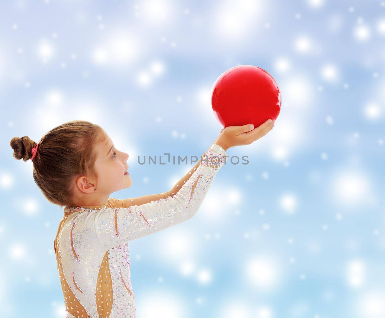 Cute little girl gymnast turned sideways to the camera , holding in his outstretched hands a red ball.On a blue background with large, white, Christmas or new year's snowflakes.