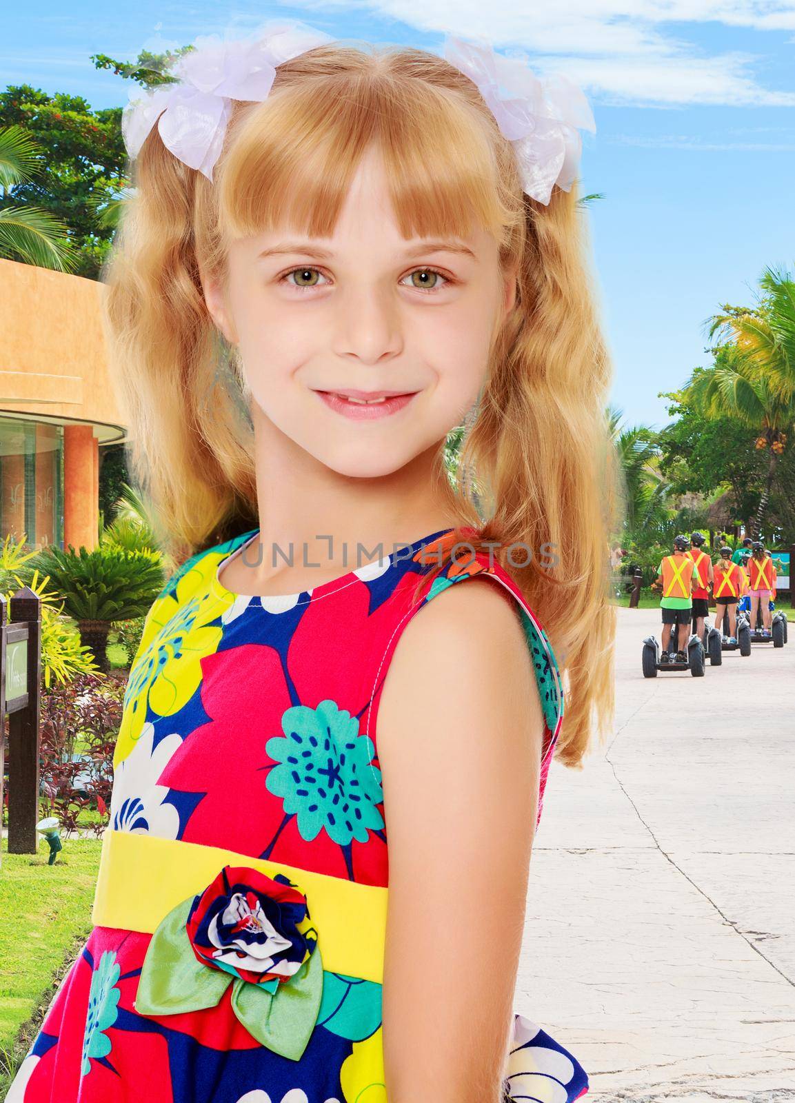 Sweet, adorable little girl with long blonde ponytails on her head tied with white bows. Close-up.On the background of the road, palm trees and blue sky with clouds.