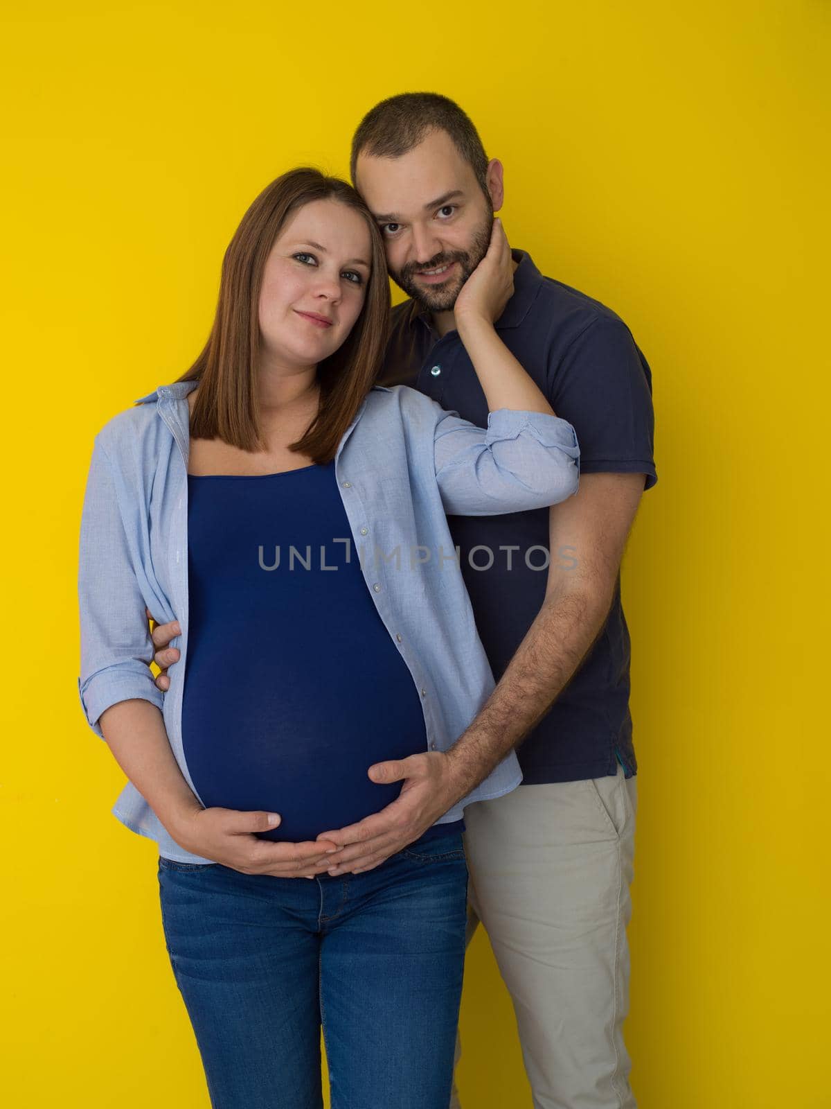 Portrait of a happy young couple,man holding his pregnant wife belly isolated over yellow background
