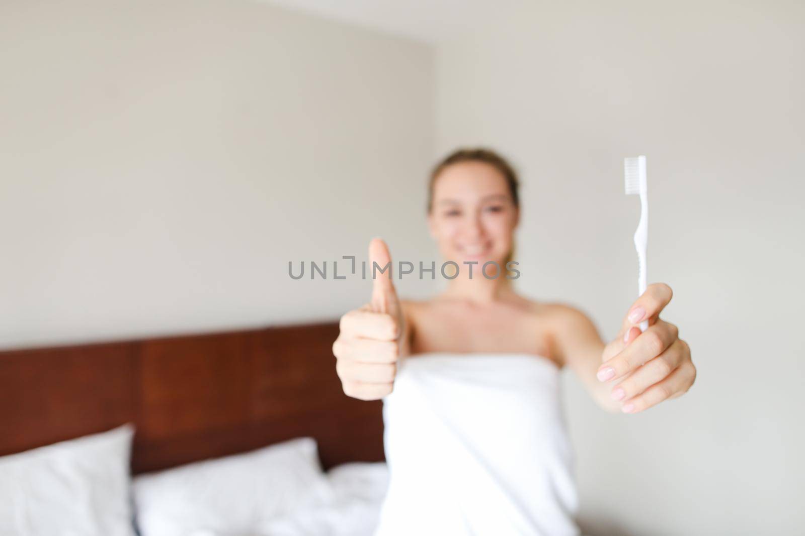Young smiling female person wrapped in white towel standing with toothbrush near bed, showing thumbs up. Concept of morning hygiene and healthy way of life.