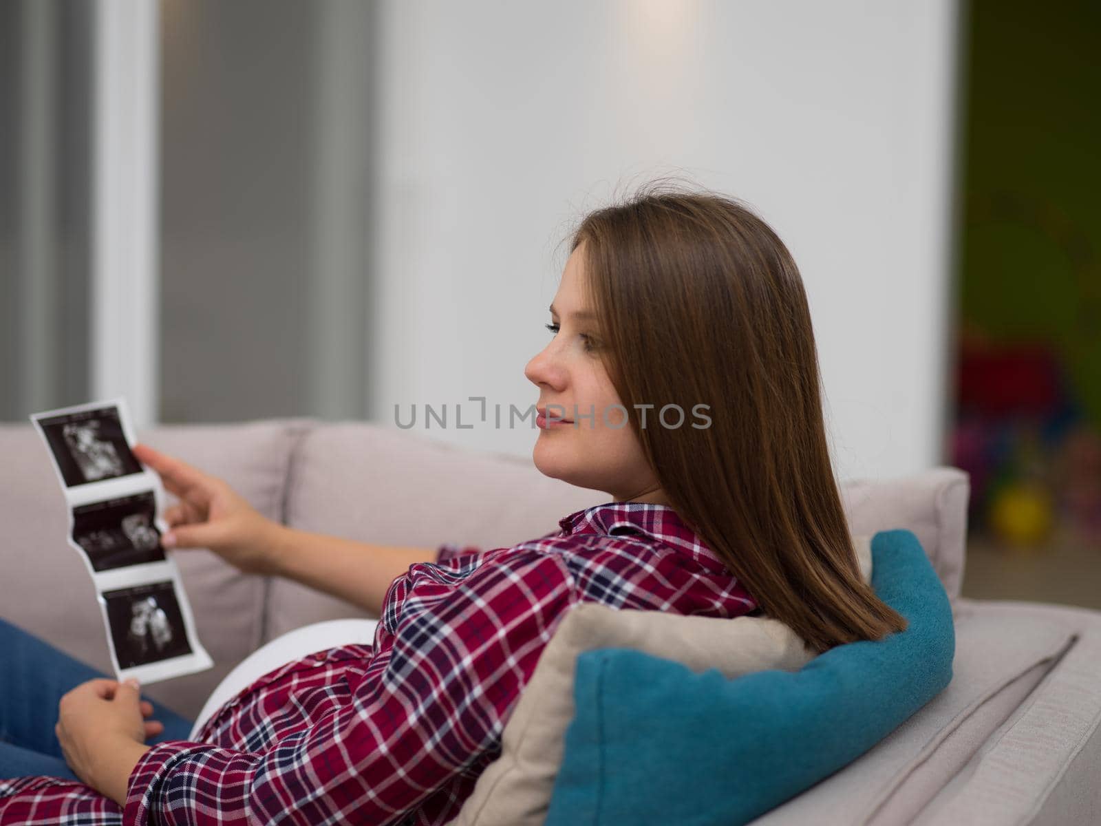 Young pregnant woman looking baby's ultrasound photo while relaxing on sofa at home