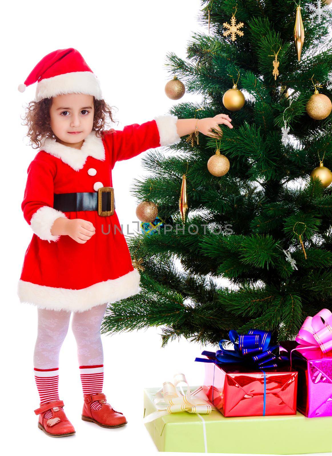 Adorable little curly-haired girl, dressed as Santa Claus decorates a Christmas tree toys.Isolated on white background.