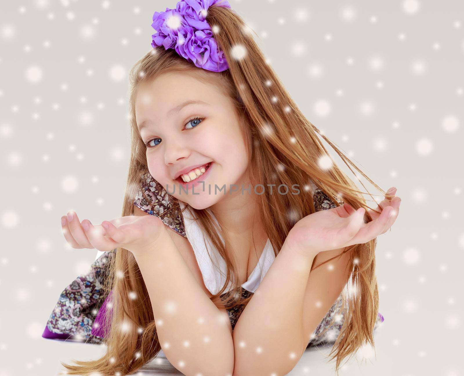 Happy little girl with a big purple bow on her head lying on the floor. Girl throws hands and smiling sweetly at the camera.On the Christmas background with white snowflakes.