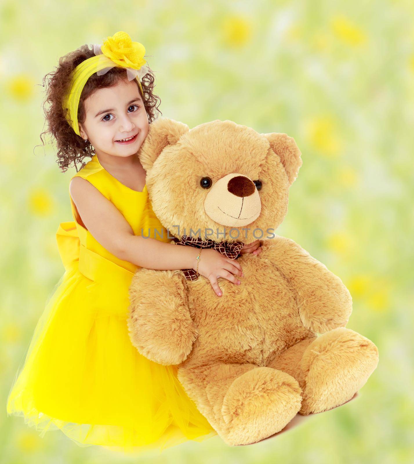 Joyful little girl in a yellow dress and bow on her head sitting on the floor. Girl hugging a big Teddy bear.Bright,floral yellow-green blurred background.