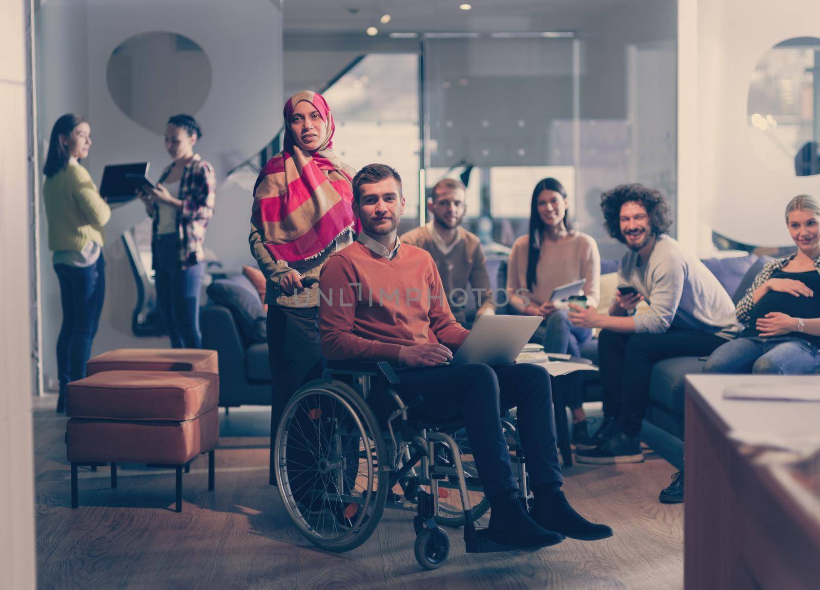 Disabled businessman in a wheelchair at work in modern open space coworking office on team meeting or brainstorming. Muslim businesswoman with hijab is assisting and helping on the project. Effective teamwork of volunteers concept in a startup business. A pregnant female coworker in the background. 