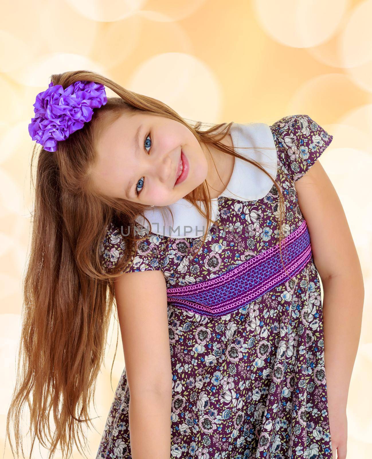 Happy little girl with a big purple bow on her head , and fancy dress. The girl bent her head to see her long beautiful hair.On a brown blurred background with white snowflakes.