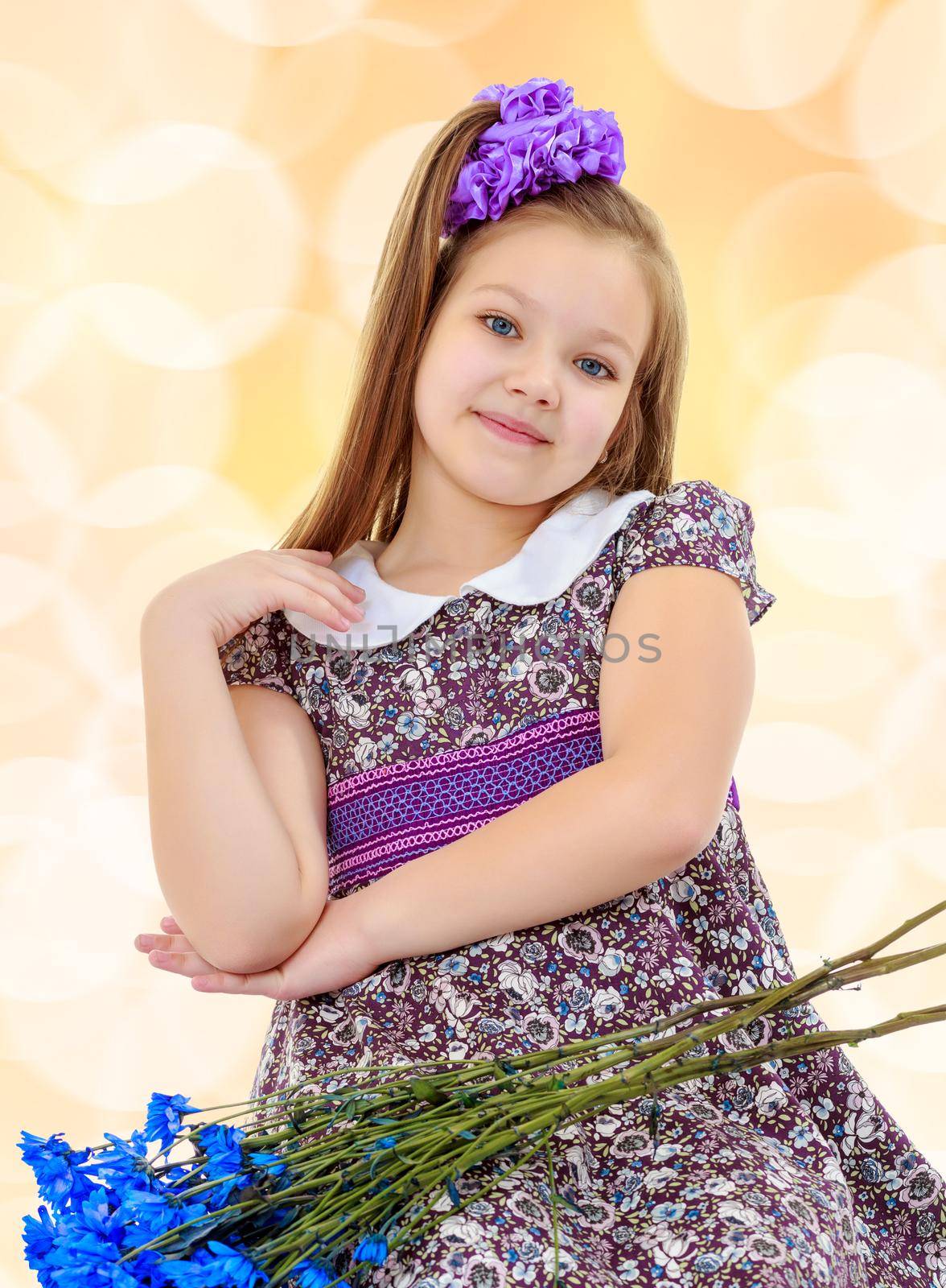 Happy little girl with a big purple bow on her head , and fancy dress. In the lap of the girls lay a bouquet of blue flowers.On a brown blurred background with white snowflakes.