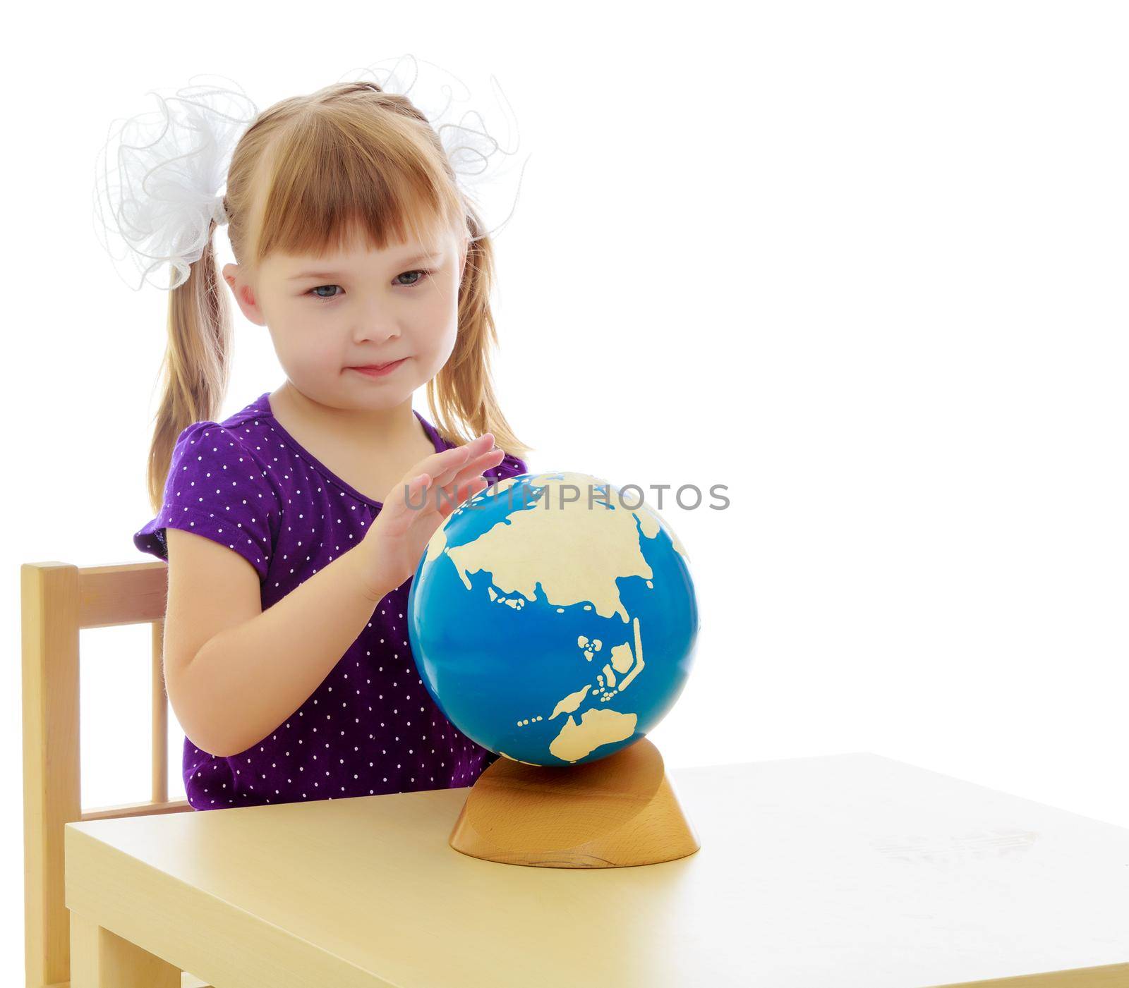 Nice little blonde girl in a blue dress. Girl sitting at table and looking at a globe.Isolated on white background.