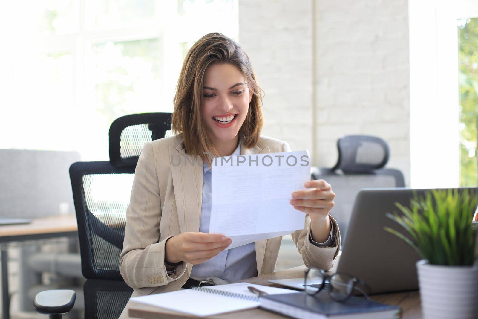 Attractive cheerful business woman checking paper documents in office, working on laptop. by tsyhun