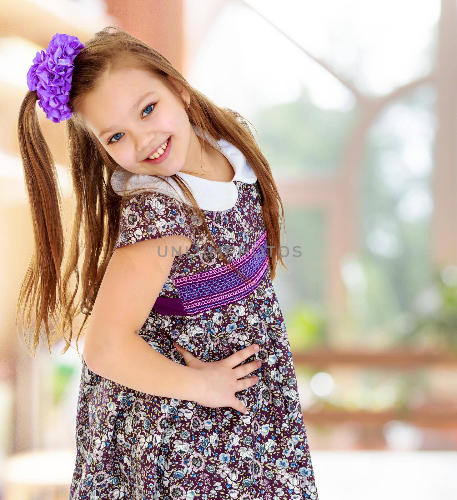 Happy little girl with a big purple bow on her head , and fancy dress. The girl bent her head to see her long beautiful hair.In the room with large, bright Windows.