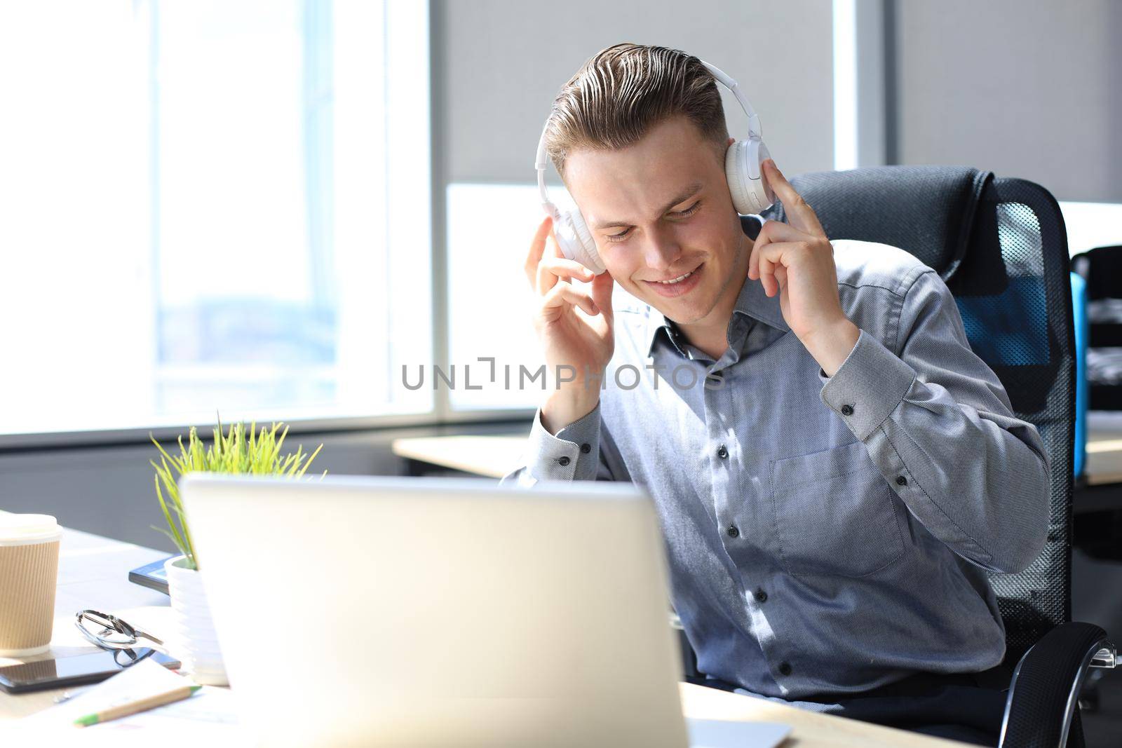 Smiling handsome businessman relaxing and listening music in earphones in a modern office.