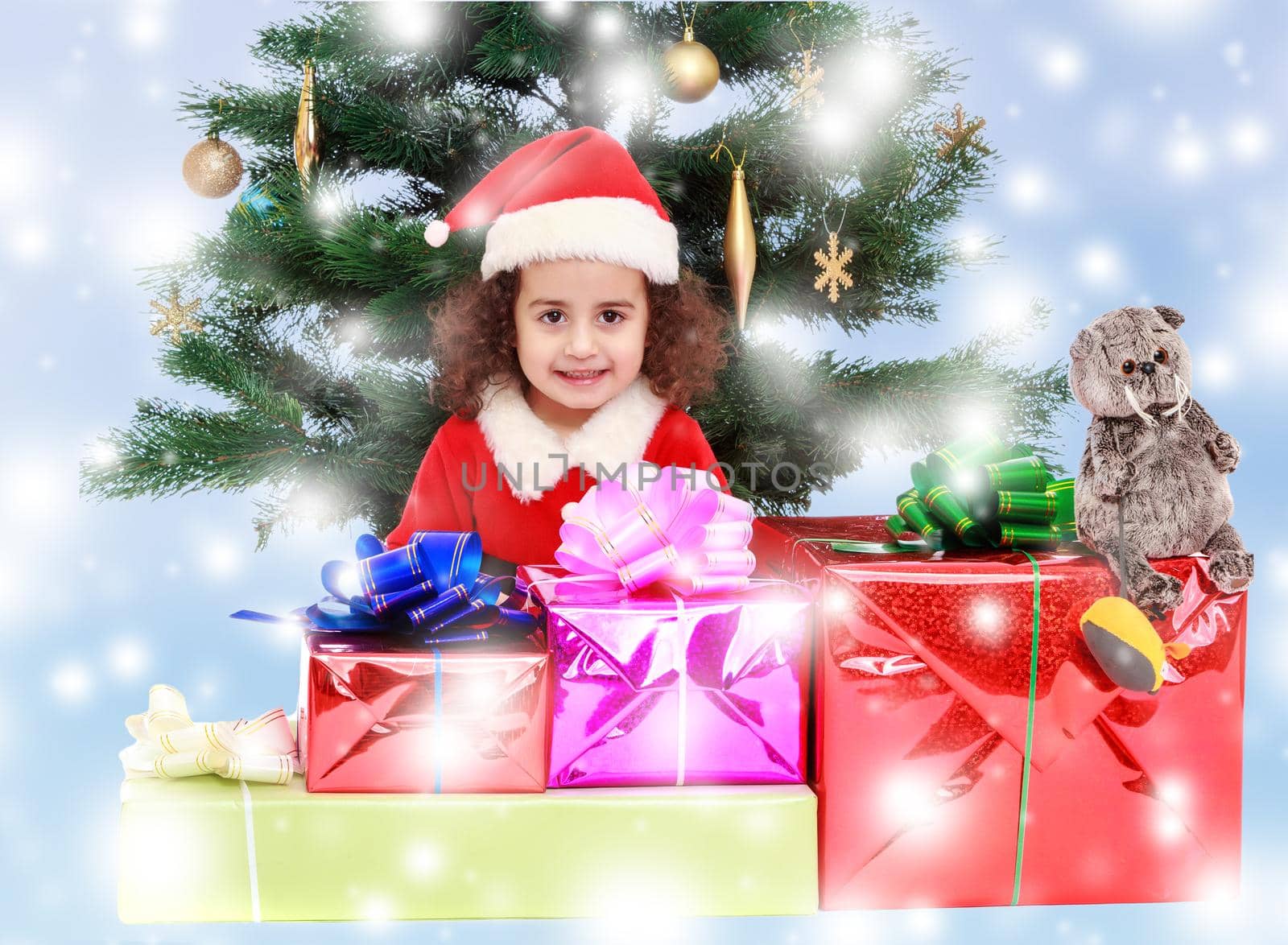 The beautiful, curly little girl in a suit and hat of Santa Claus sits near Christmas tree with bunch of gifts.Blue winter background with white snowflakes.