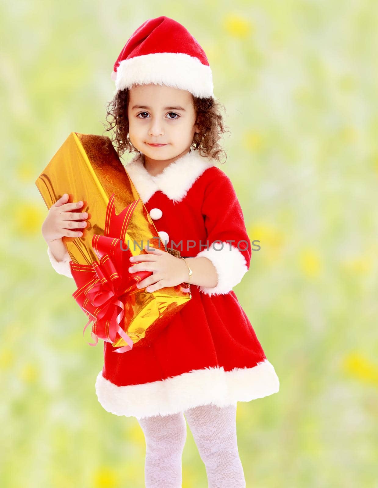 Cute little curly girl in a coat and hat of Santa Claus holding a box with a gift.Bright,floral yellow-green blurred background.