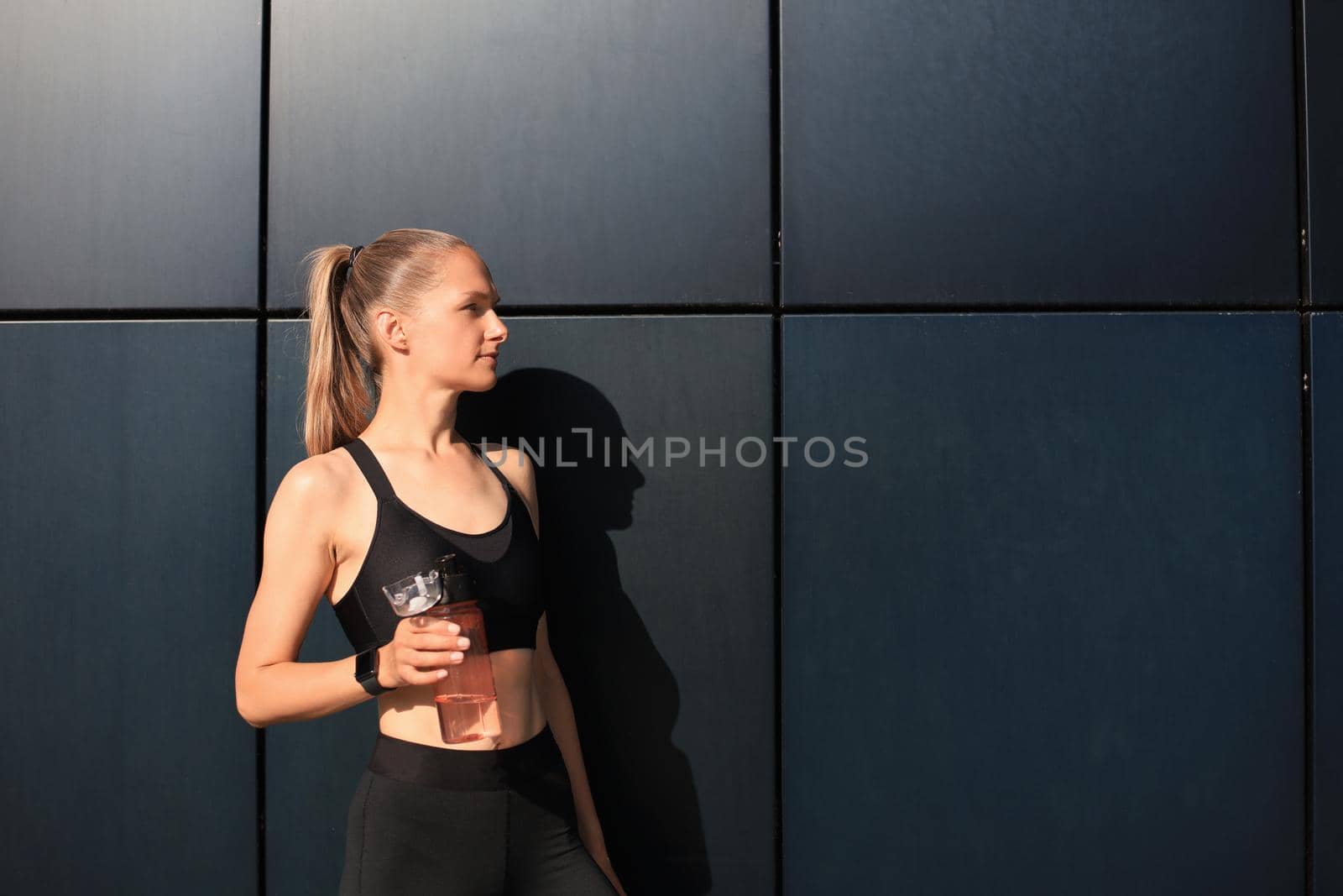 Beautiful female runner is standing outdoors holding water bottle. Fitness woman takes a break after running workout